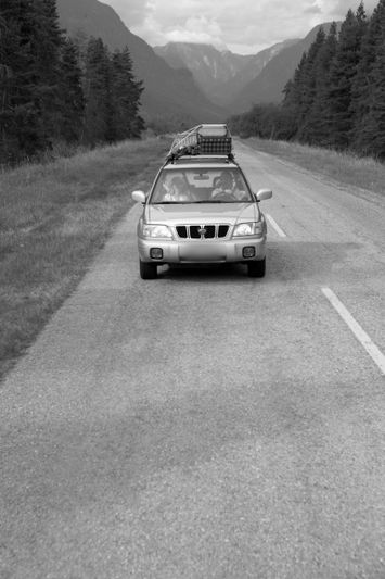 Foto en blanco y negro de formato estrecho que muestra un coche todoterreno con equipaje en su techo, circulando por una carretera asfaltada en medio del bosque, con un paisaje montañoso de fondo. La foto ha sido sometida a un proceso de anonimización de la matrícula.