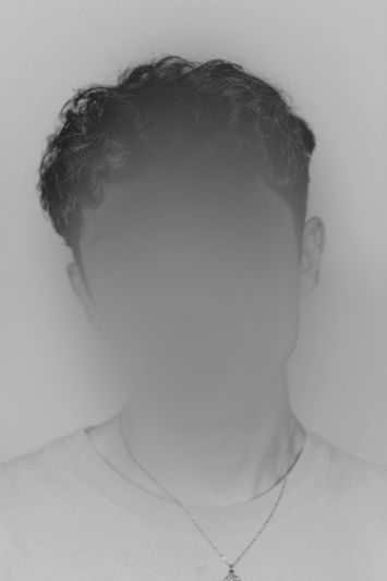 Czarno-białe portret osoby o kręconych włosach; rysy twarzy zamazane gładką mgiełką, ubrana w koszulkę i naszyjnik.