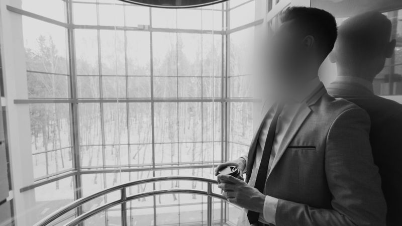 A person in a suit stands in a glass elevator, looking out at a snowy landscape. The face is blurred, reflecting on the glass.