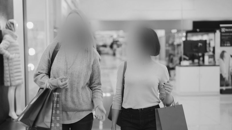 Deux personnes aux visages flous, portant des sacs de courses, traversent un centre commercial. À l'arrière-plan, des mannequins présentent des vêtements. Image en noir et blanc.