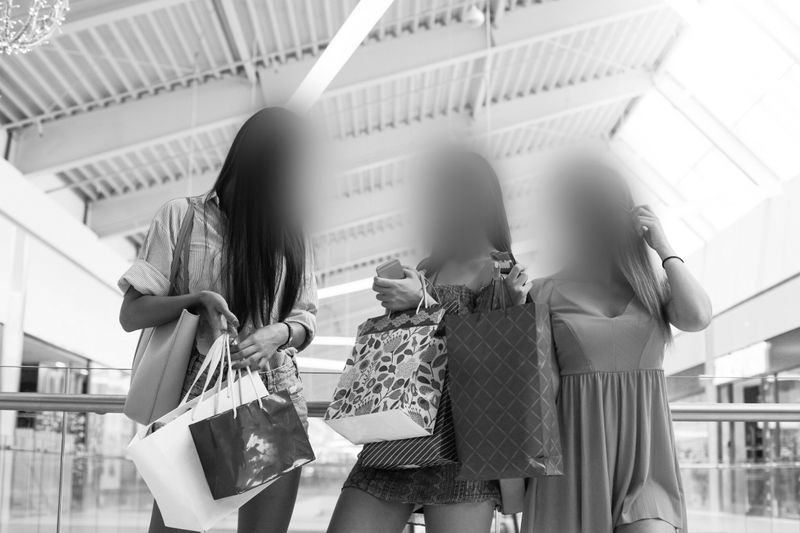 Trois personnes dans un centre commercial, tenant des sacs de courses, le visage flouté, debout sous de hauts plafonds baignés de lumière naturelle. Image en noir et blanc.