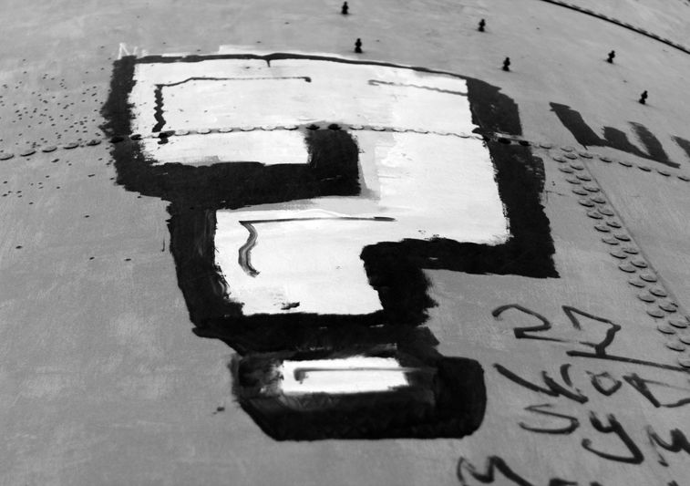Black and white image of a bold, painted question mark on a metal surface, with some handwritten text and rivets visible.