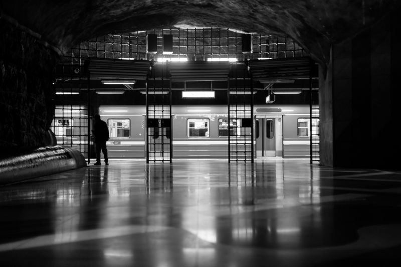 Osoba stoi na słabo oświetlonej stacji metra, w tle przejeżdża pociąg. Scena jest czarno-biała.