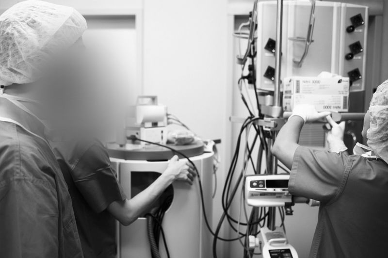 Two medical professionals in a hospital setting, adjusting equipment and monitoring devices, with both wearing scrubs and caps.
