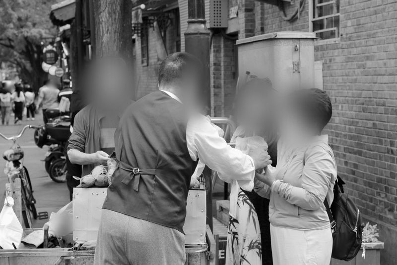 Trois personnes à l'intérieur d'une voiture, deux sur les sièges avant et une penchée à l'intérieur depuis l'extérieur, les visages flous. Image en noir et blanc.