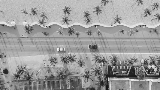 Widok z lotu ptaka na drogę wzdłuż plaży, z palmami rzucającymi długie cienie, jadącym samochodem i pobliskimi budynkami.