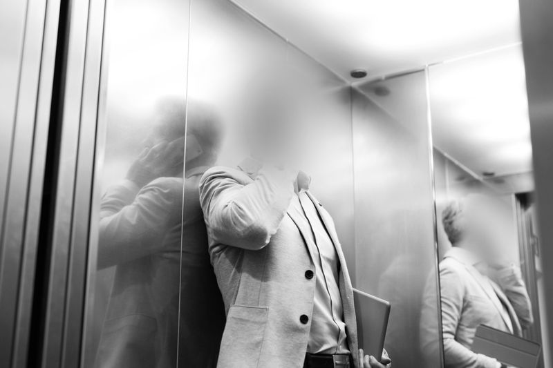Man in a suit leaning against elevator wall, face obscured. A laptop is under his arm, and reflections are visible on the metal surface. Black and white.