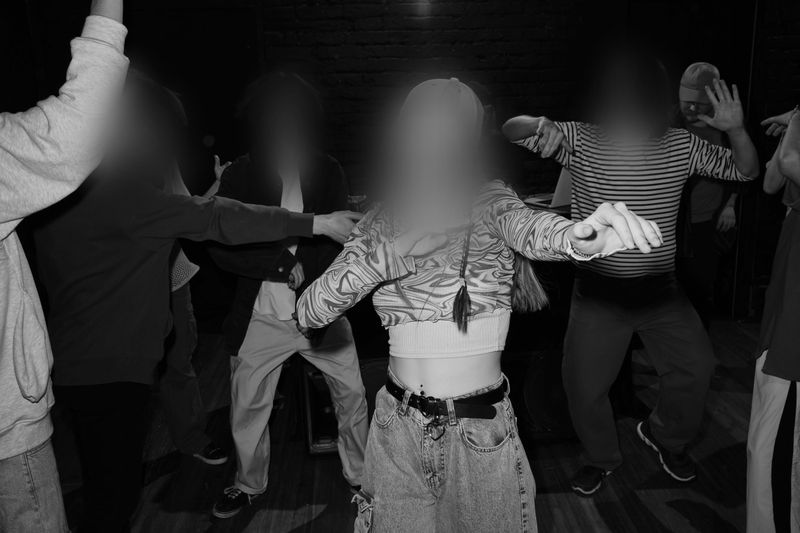 A group of people dancing energetically in a dimly lit room, with motion blur and indistinct faces, creating a lively atmosphere.