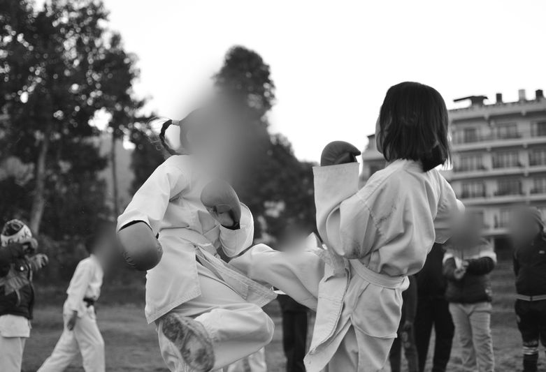 Two kids in martial arts uniforms practice sparring outdoors, surrounded by onlookers and trees, in a black and white photo.
