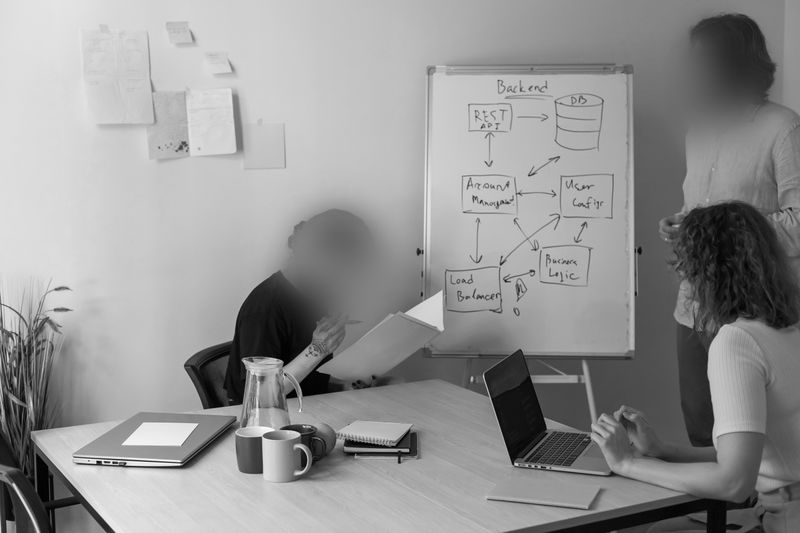 Three people in a meeting room at a table with laptops and notes, discussing a diagram on a whiteboard about backend processes.