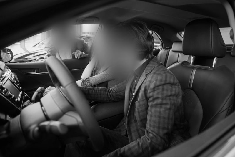 Trois personnes à l'intérieur d'une voiture, deux sur les sièges avant et une penchée à l'intérieur depuis l'extérieur, les visages flous. Image en noir et blanc.