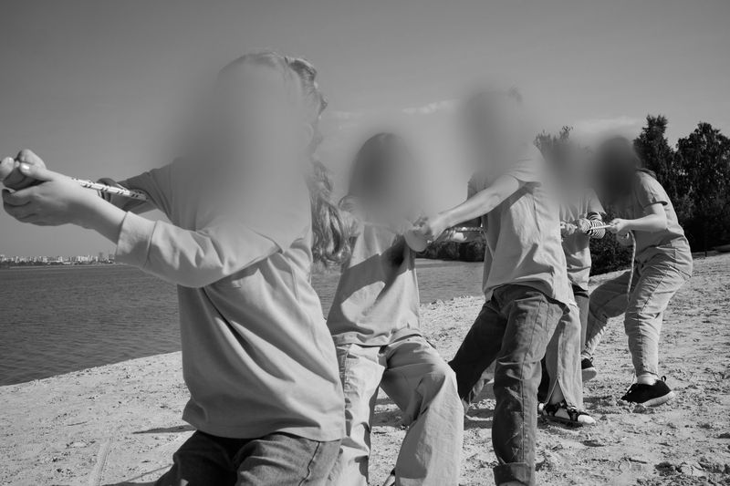 Group of people engaged in a tug-of-war on a sandy beach near a body of water, with blurred faces and trees in the background.