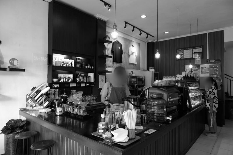 Photo en noir et blanc d'un café chaleureux où l'on voit deux personnes derrière le comptoir, entourées de machines à café et d'étagères remplies de produits.