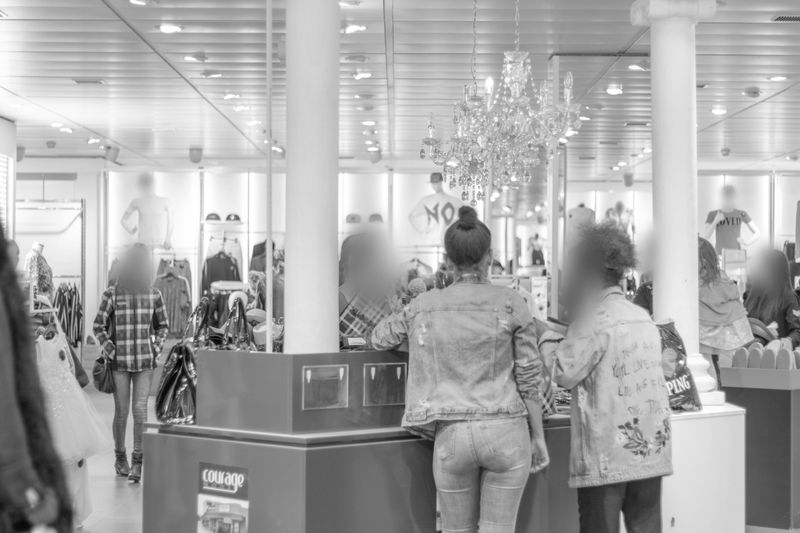 Des clients faisant leurs achats dans un magasin de vêtements bien éclairé, avec un lustre, des portants remplis de vêtements et une caisse.