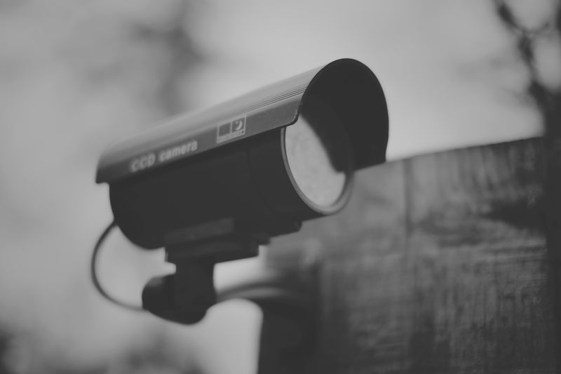 Image en noir et blanc d'une caméra de vidéosurveillance montée sur un poteau en bois, tournée vers la gauche. Arrière-plan flou.