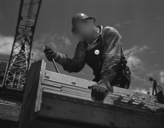 Un ouvrier du bâtiment en équipement de protection utilise des machines sur une structure en acier, avec une grue et un ciel bleu en arrière-plan.