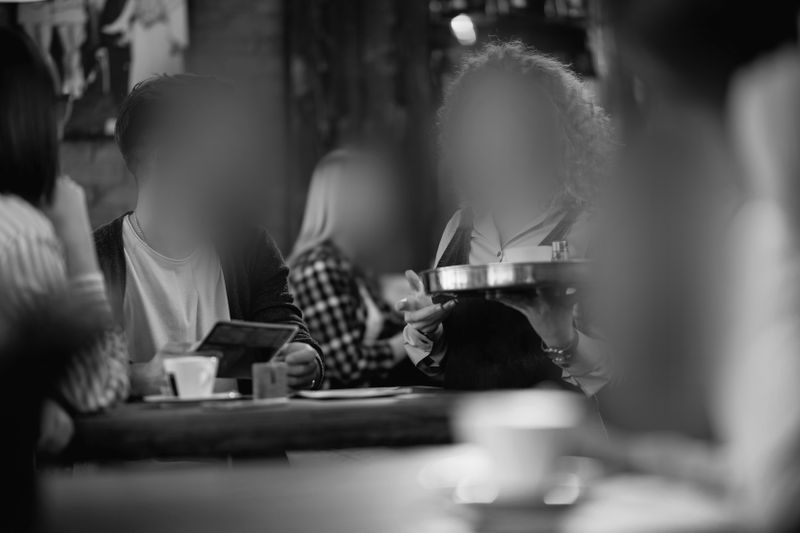 Image floue en noir et blanc montrant des personnes assises en train de discuter dans un café, avec un serveur portant un plateau.