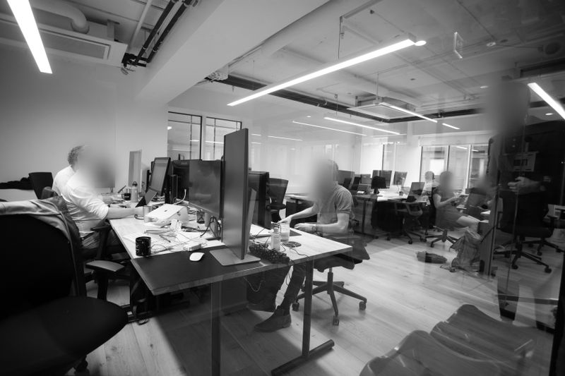 Black and white image of an open-plan office with people working at desks, computers, and modern lighting overhead.