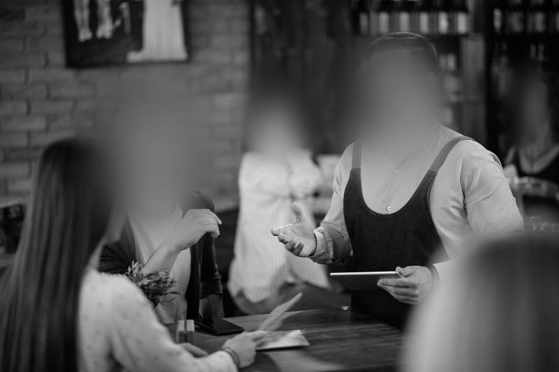 Un groupe de personnes, dont les visages sont flous, est réuni autour d'une table dans un café. Un serveur portant un tablier prend une commande, un bloc-notes à la main. Image en noir et blanc.