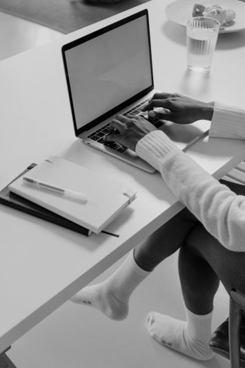 Persona escribiendo en una laptop sobre un escritorio blanco, con cuadernos, un bolígrafo y un vaso de agua cerca. Lleva suéter y calcetines. Imagen en blanco y negro.