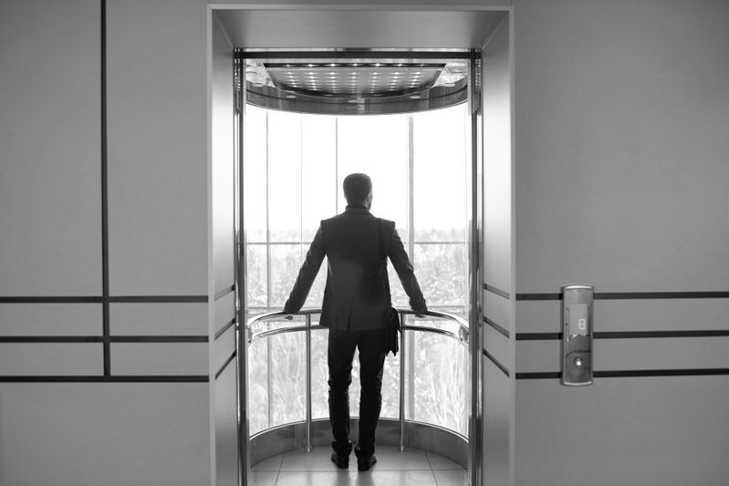 Man in a suit standing in a glass elevator, facing outward with a briefcase, overlooking a cityscape. Black and white image.