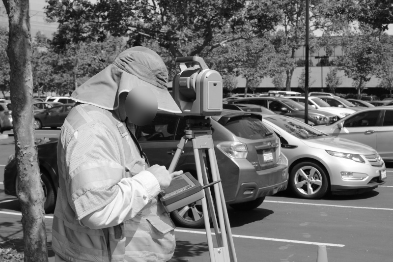 Anonymisierungsmethoden zur Verschleierung der Identität von Personen, Vermesser in Schutzkleidung verwendet Ausrüstung auf einem Parkplatz mit Bäumen und geparkten Autos im Hintergrund.