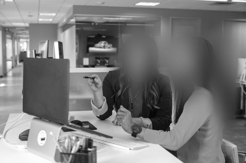Two people working at a desk in an office, discussing something on a computer screen. Office supplies are visible on the desk.