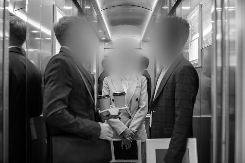Three people in business attire stand in an elevator, holding papers and coffee cups. The scene is in black and white.