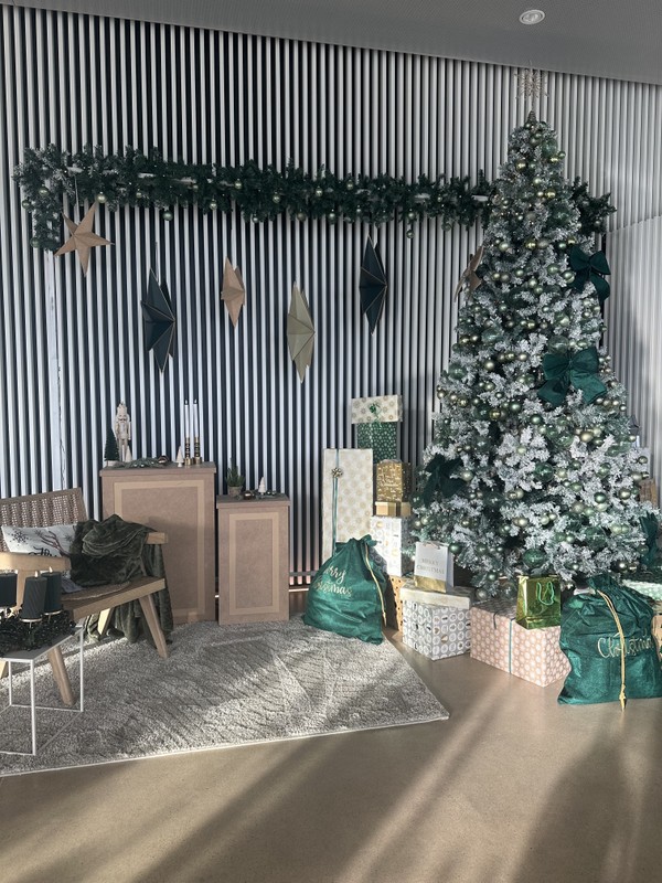 Modern Christmas setup with a tall, decorated tree, wrapped gifts, and green bags. Wooden furniture and star-shaped ornaments add to the festive decor.