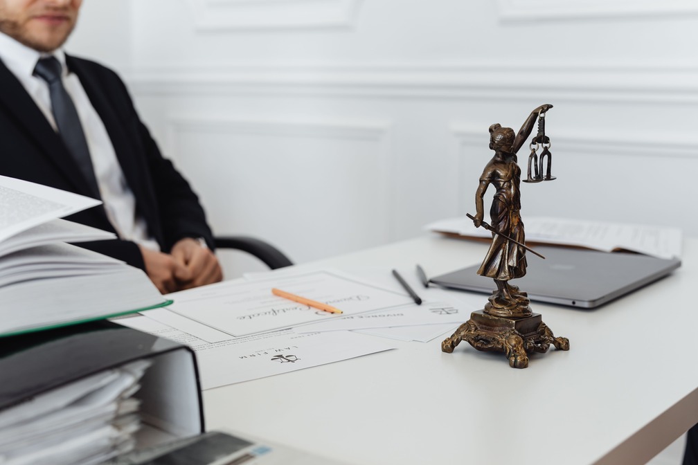 A man in a suit sits at a desk with a Lady Justice statue, documents, and a laptop in an office setting.