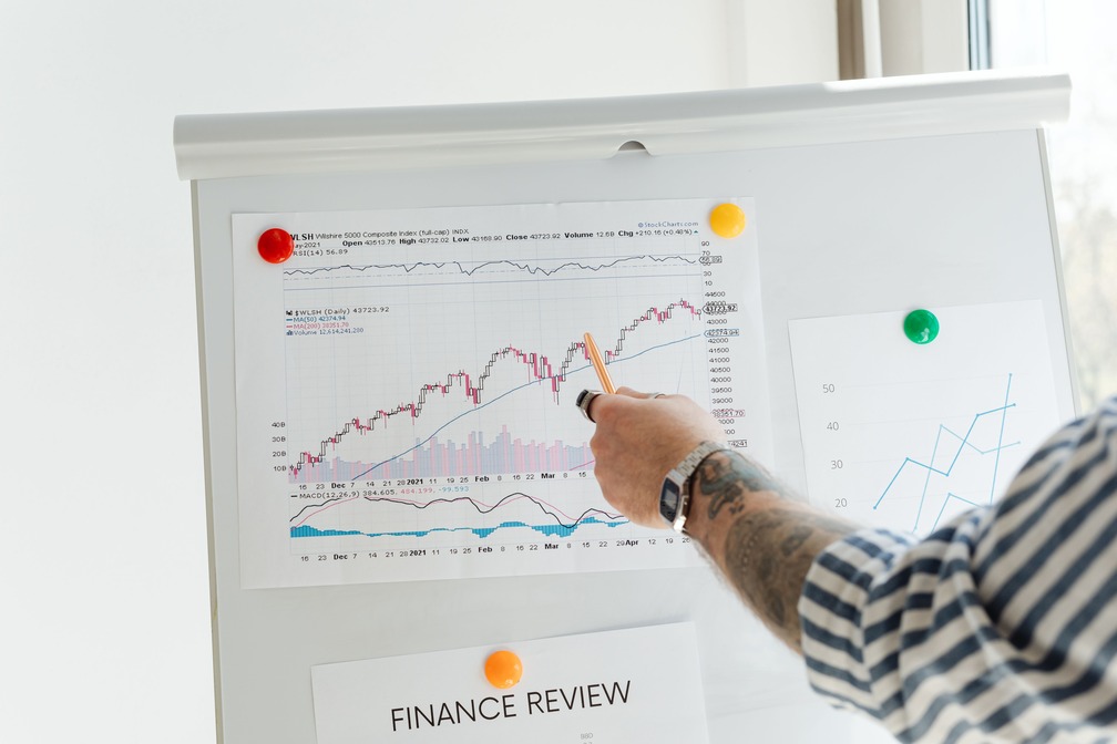 Person pointing at a line graph on a whiteboard during a finance review, with charts and graphs pinned alongside.