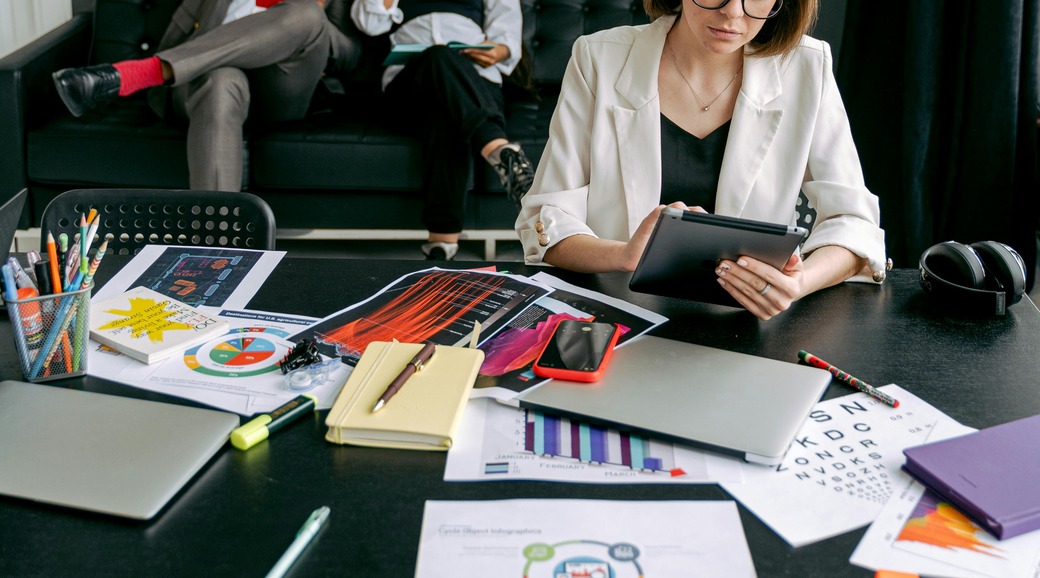 Woman in white blazer using a tablet at a cluttered desk with laptops, charts, notebooks and headphones; two people sit on a couch behind her.