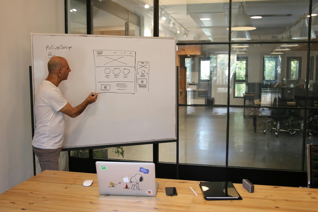 Man at a whiteboard sketches a website layout in a modern office, with a laptop and notebook on a wooden table nearby.