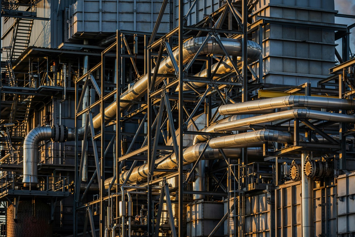 Complex network of stainless steel pipes and metal scaffolding on an industrial plant, bathed in warm late-afternoon light.