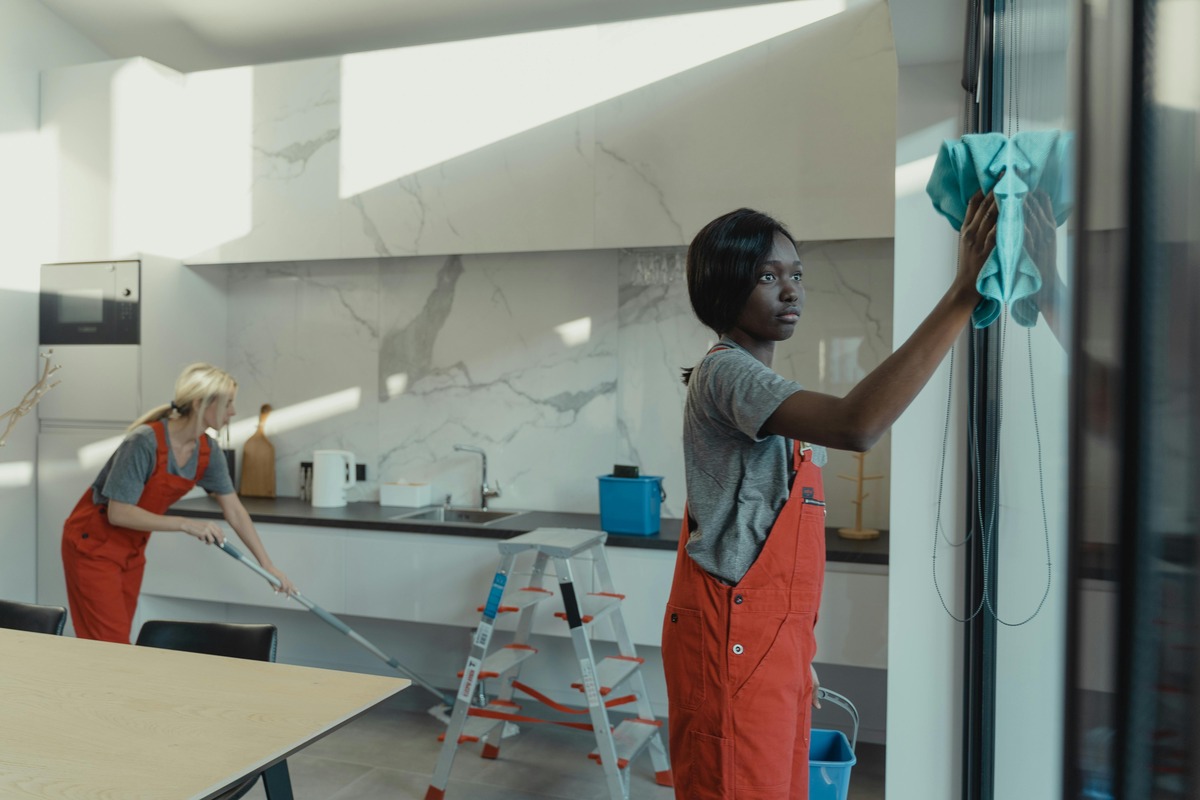 Two cleaners in orange overalls cleaning a modern kitchen; one wipes a glass door while the other mops the floor near the counter.
