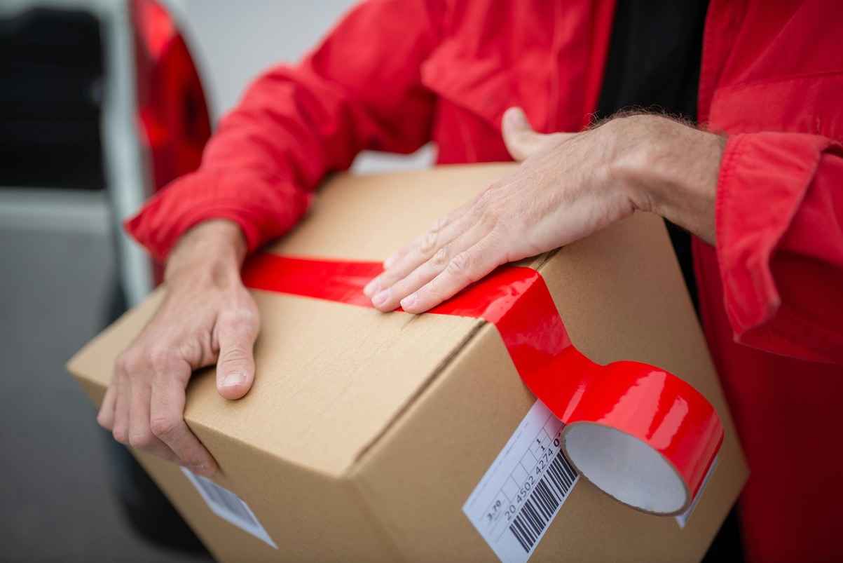 Person sealing a cardboard box with red tape, wearing a red jacket. A car is visible in the background.