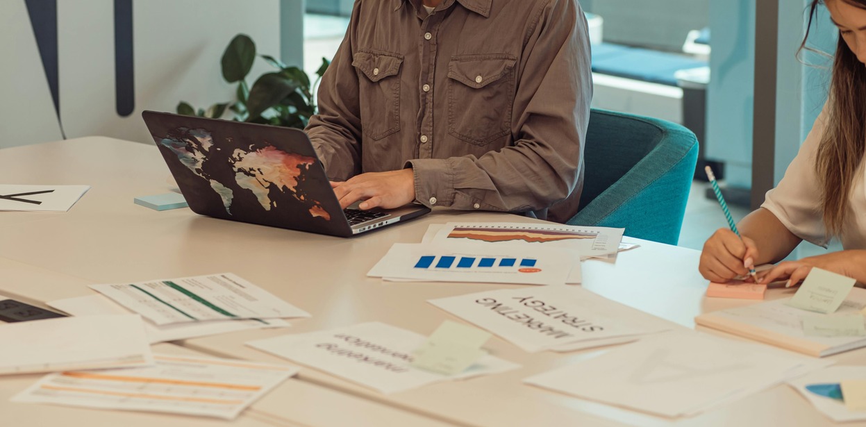 Two colleagues at a meeting table with a laptop showing a world-map sticker, charts and papers spread out as one types and another writes.