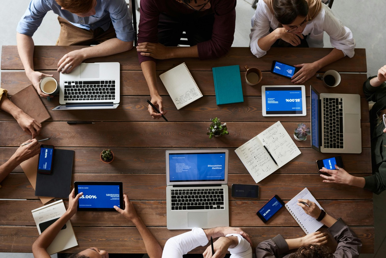 Overhead view of a collaborative meeting: diverse people around a wooden table with laptops, tablets, smartphones, notebooks, and coffee.