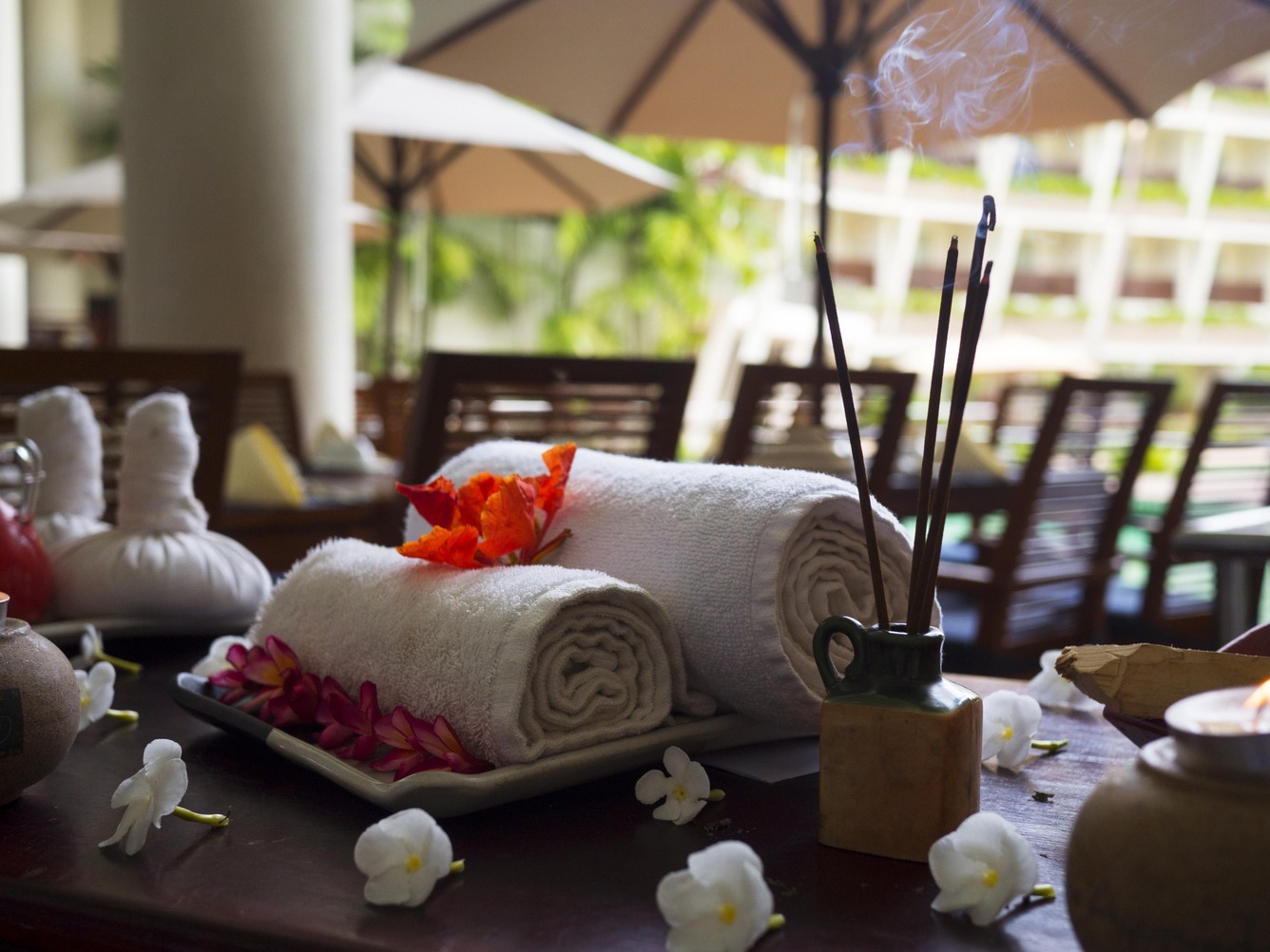 Rolled white towels with orange flowers in a spa salon, surrounded by orchids on a table, incense burning nearby, creating a serene spa ambiance.