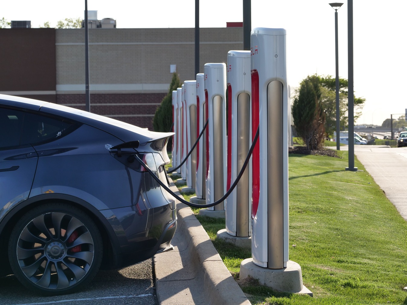 An electric car is parked and charging at a row of charging stations in a parking lot, with a modern building in the background.