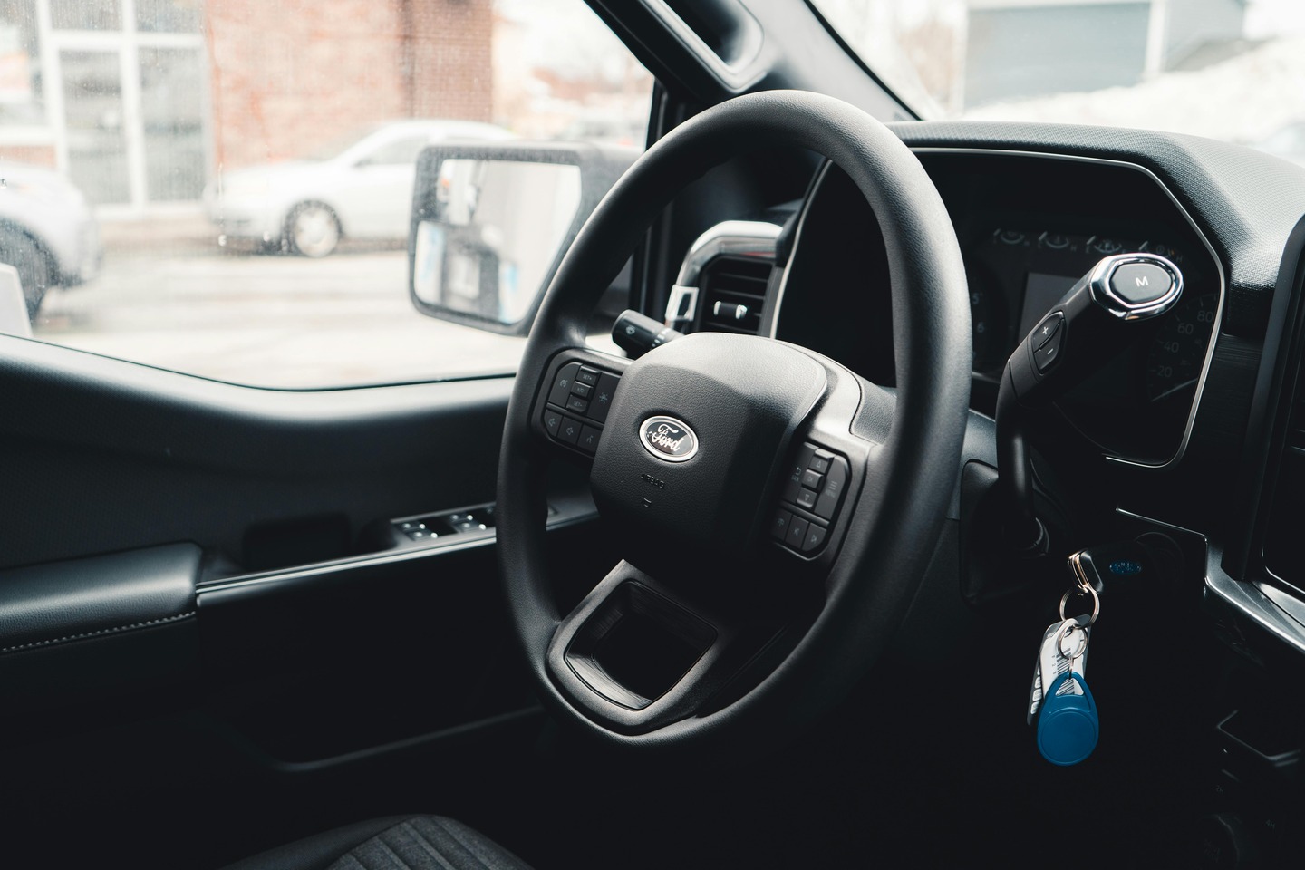 Interior view of a modern Ford truck steering wheel with ignition key fob hanging and a street visible through the window.