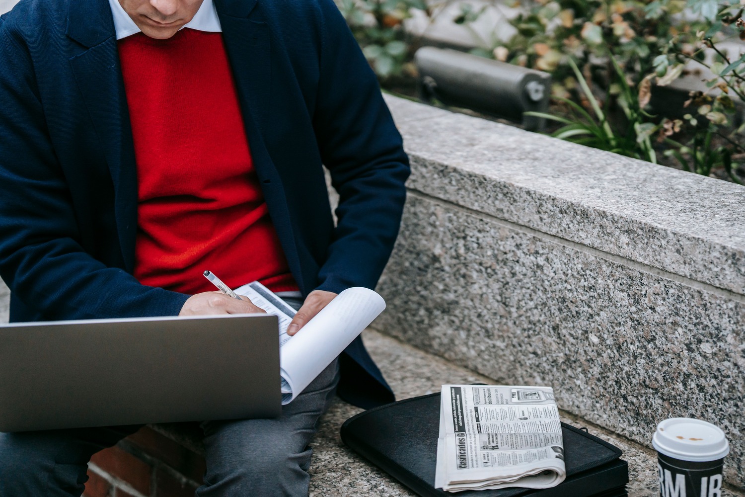 Man in a red sweater and blue jacket writing on a notepad, with a laptop on his lap. A newspaper and coffee cup sit on a stone bench beside him.