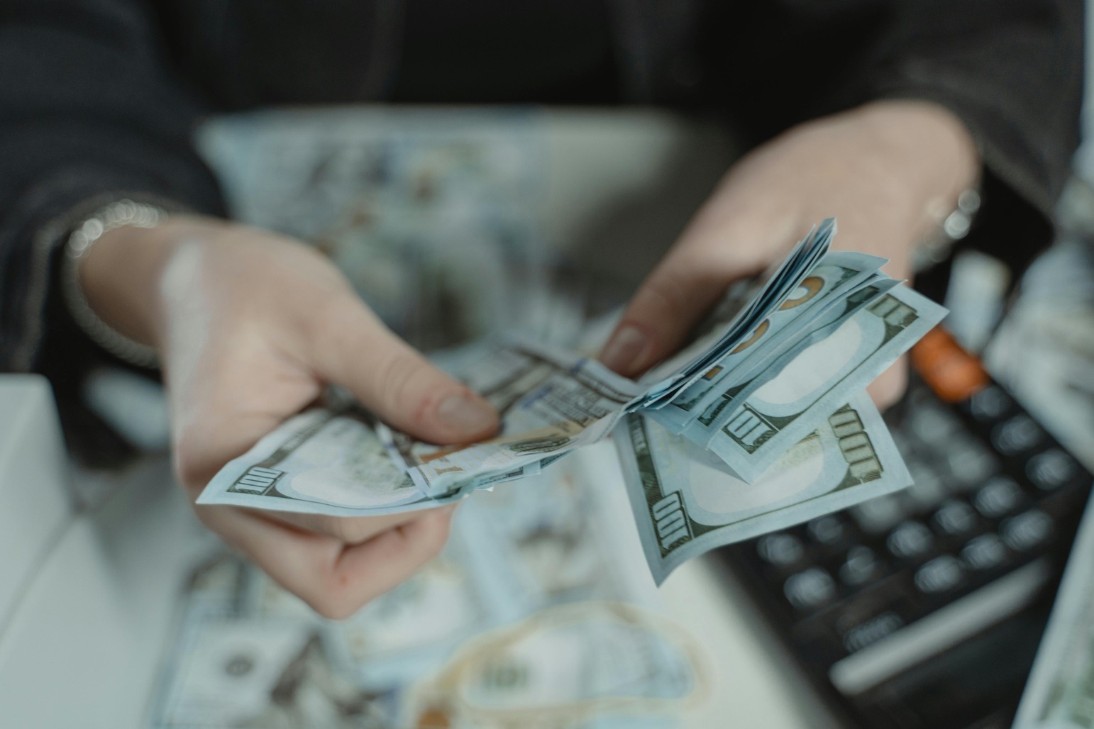 Person counting a stack of U.S. hundred-dollar bills with a calculator in the background, representing revenue or financial management.