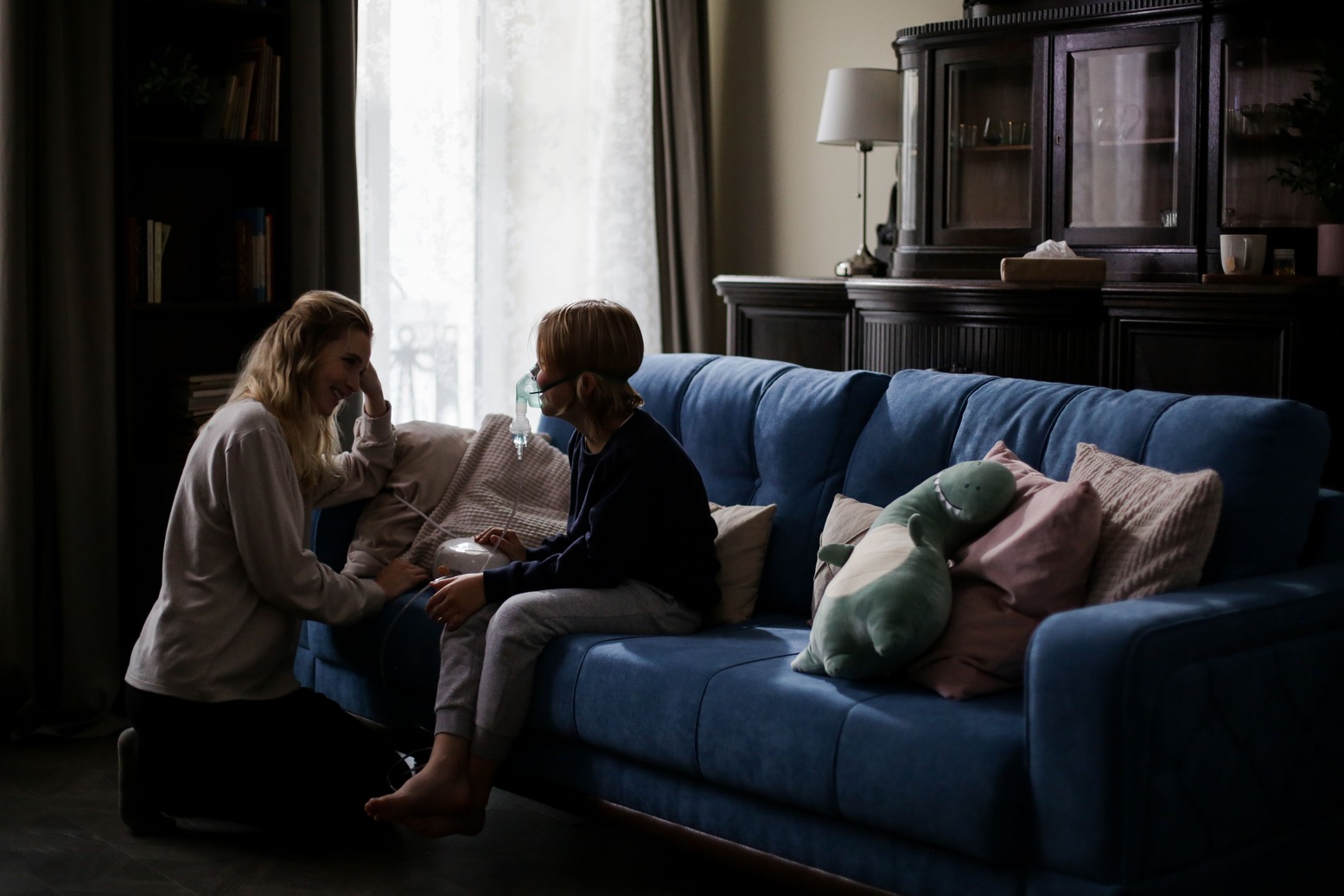 A woman kneels beside a child wearing a mask seated on a blue sofa in a dimly lit room, conveying a comforting and caring interaction.