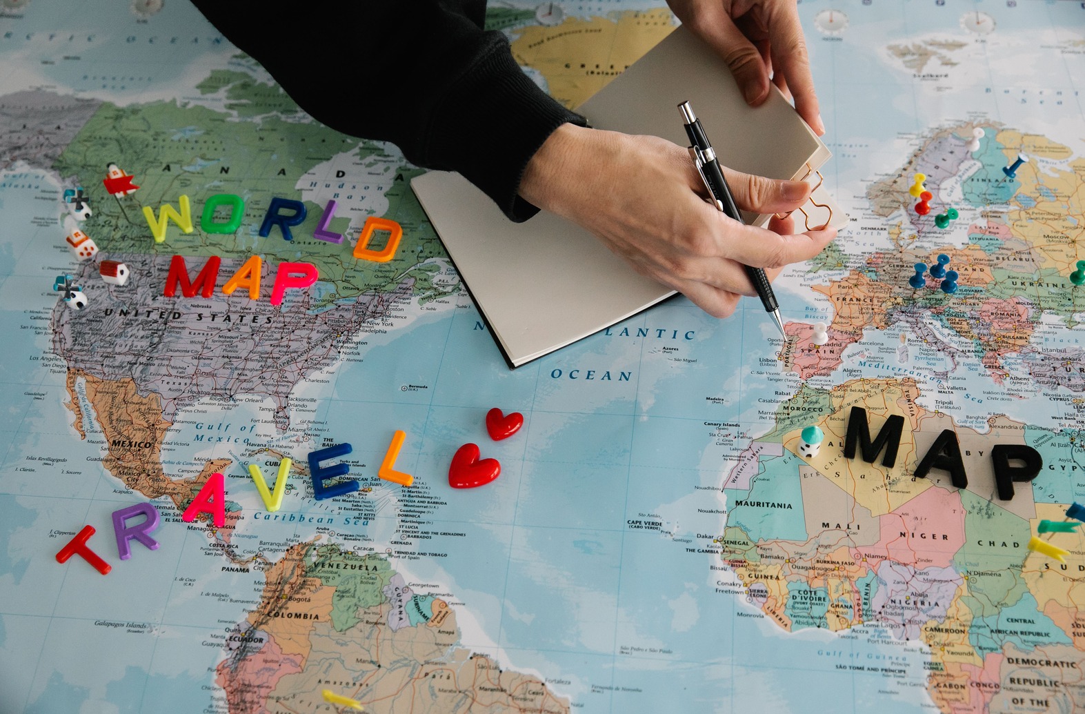 Hands holding a pen over a notebook on a world map, with colorful letters spelling "World Map" and "Travel," and pushpins marking locations.