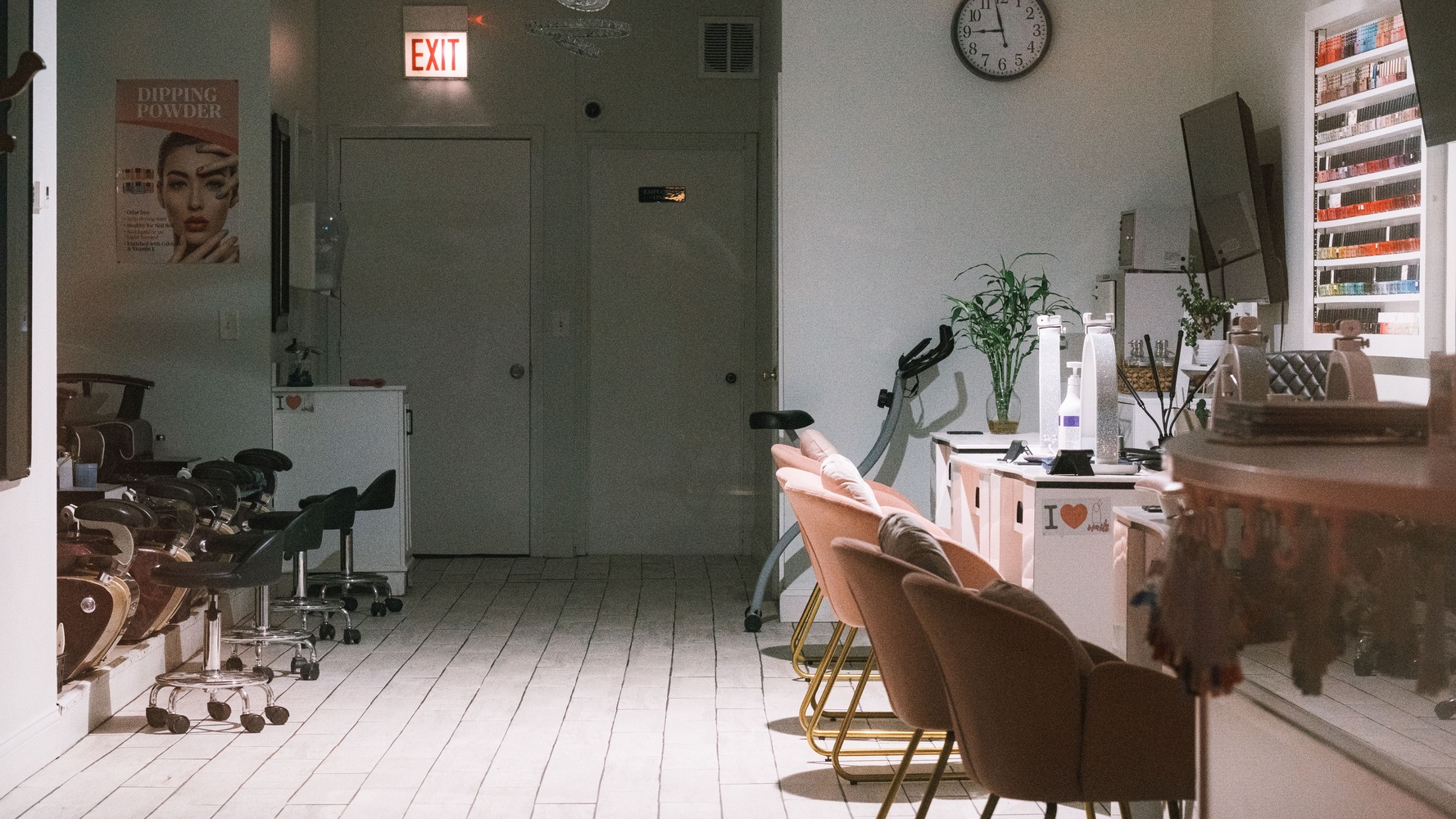 A dimly lit nail salon with beige chairs, manicure tables, a wall clock, and an exit sign.