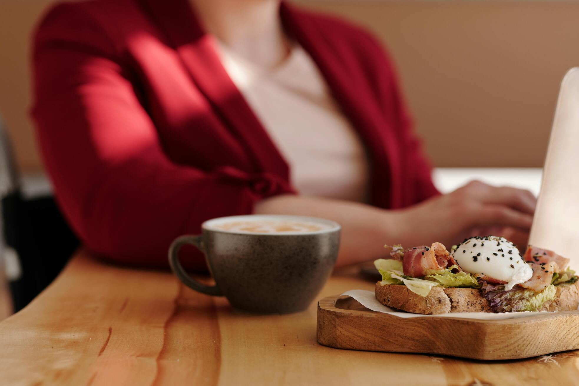 A person in a red jacket sits at a wooden table with a cappuccino and a plate of avocado toast topped with poached egg and sesame seeds.