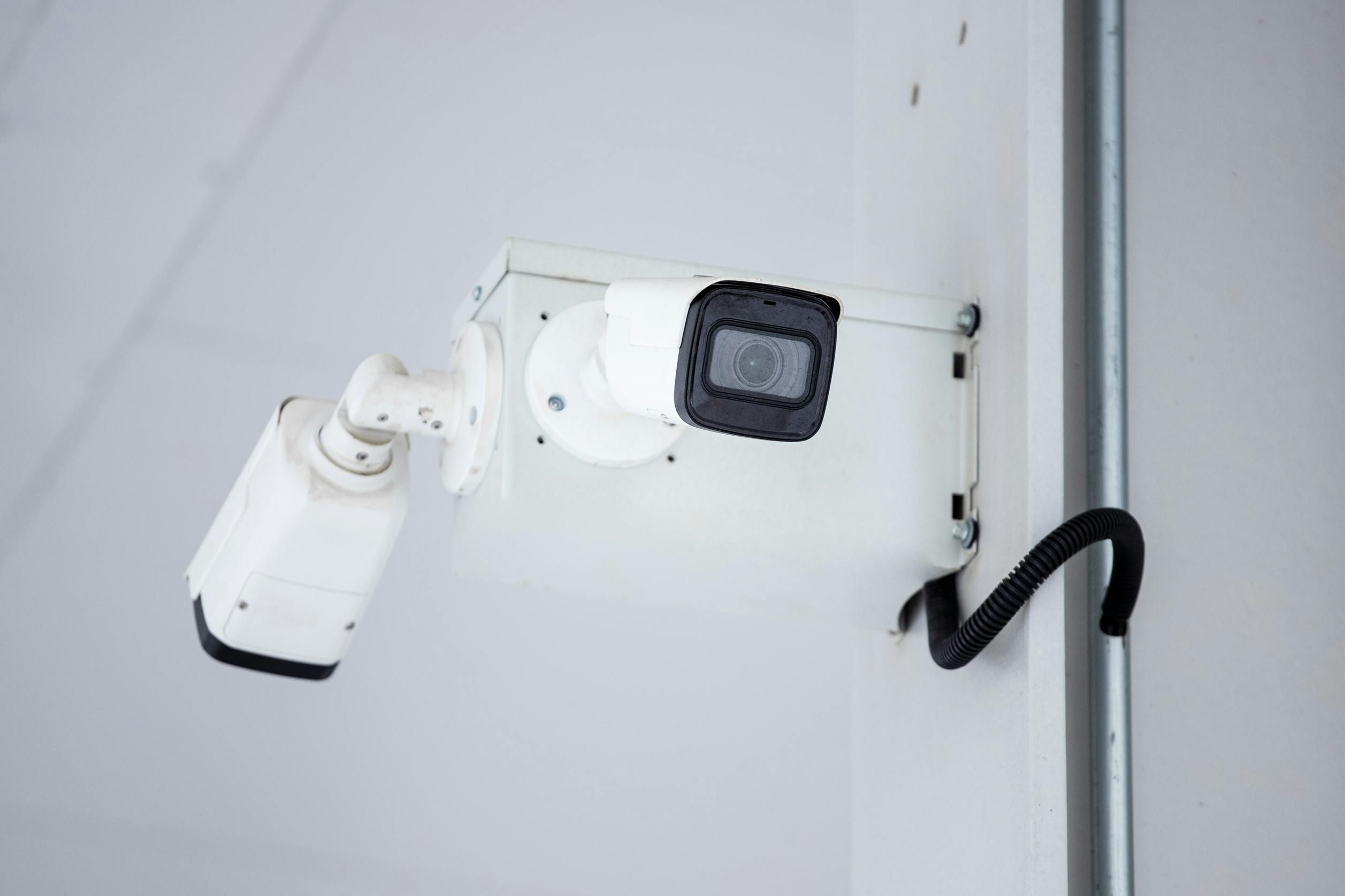 A white security camera mounted on a wall, angled downward, with a black cable extending from it.