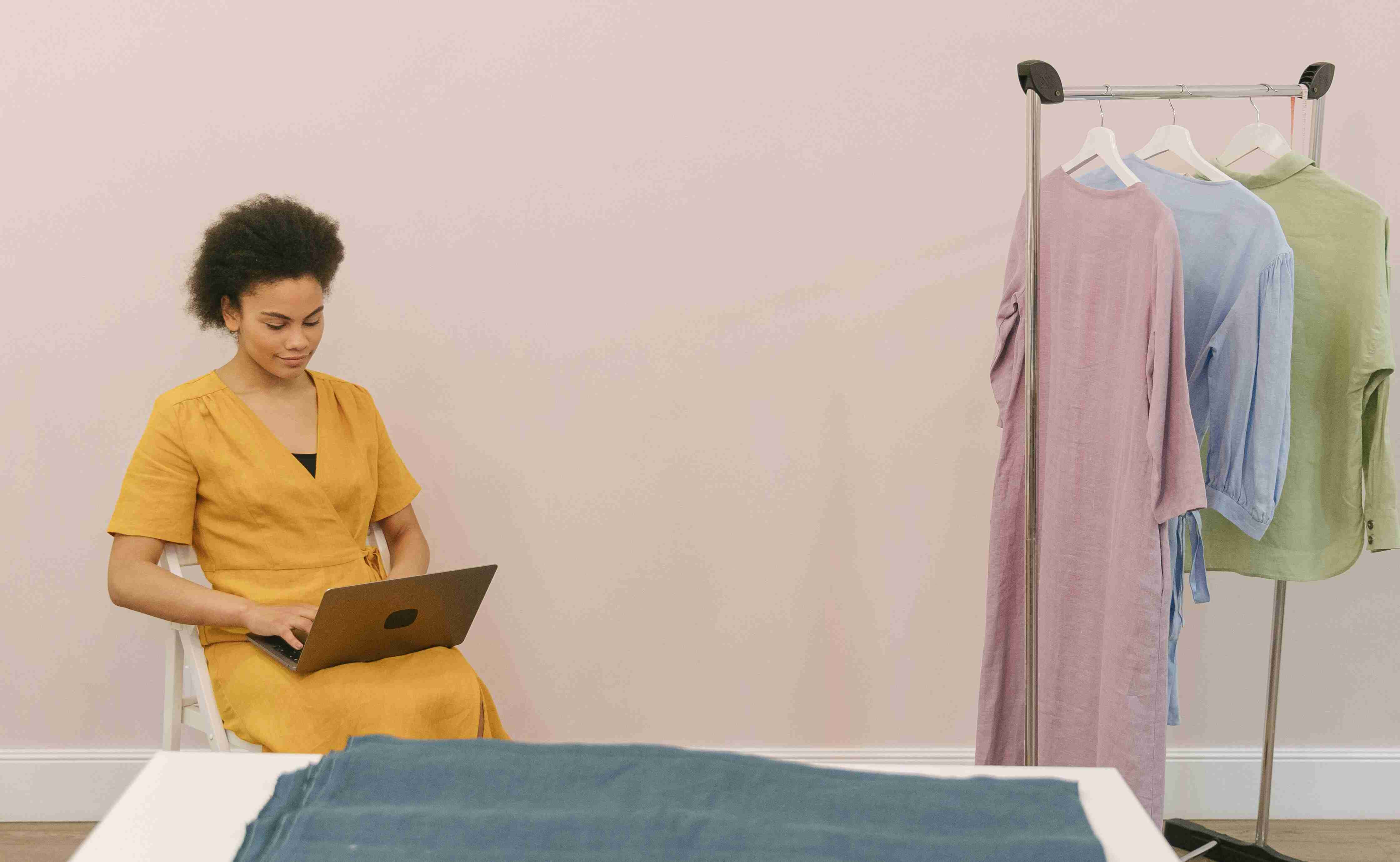 Woman in a yellow dress works on a laptop, sitting near a rack with pastel-colored dresses, in a minimalist room.