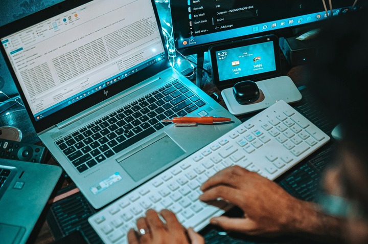 Person's hands typing on a white keyboard beside a laptop and multiple monitors showing data.